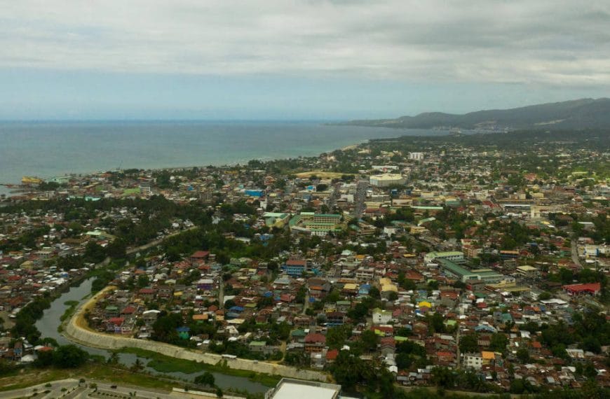 Aerial view of Iligan City with densely packed buildings, a winding river, and a mountainous backdrop.