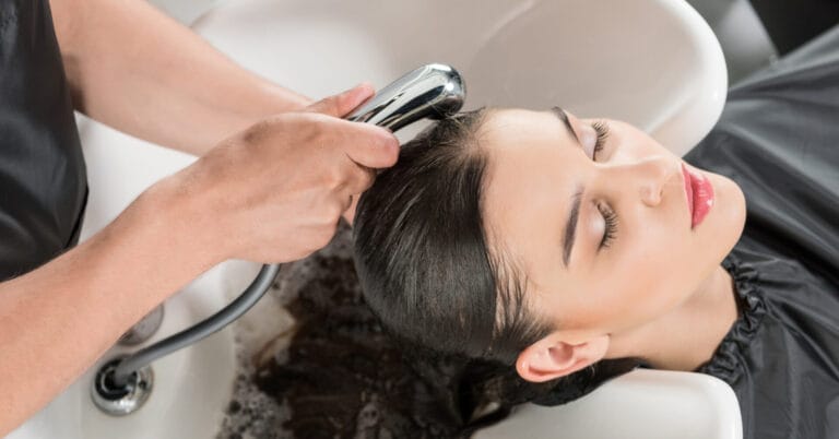 person with eyes closed getting hair washed at a quality grooming salon, water flowing from a hand held nozzle.