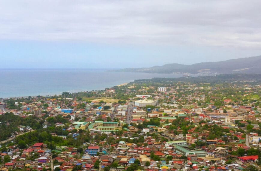 aerial view of a coastal iligan city with colorful rooftops, lush greenery, and hills under a cloudy sky.