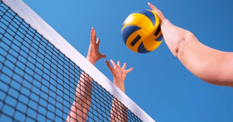 two people reach for a yellow and blue volleyball above a net at the diyandi festival under a clear blue sky.