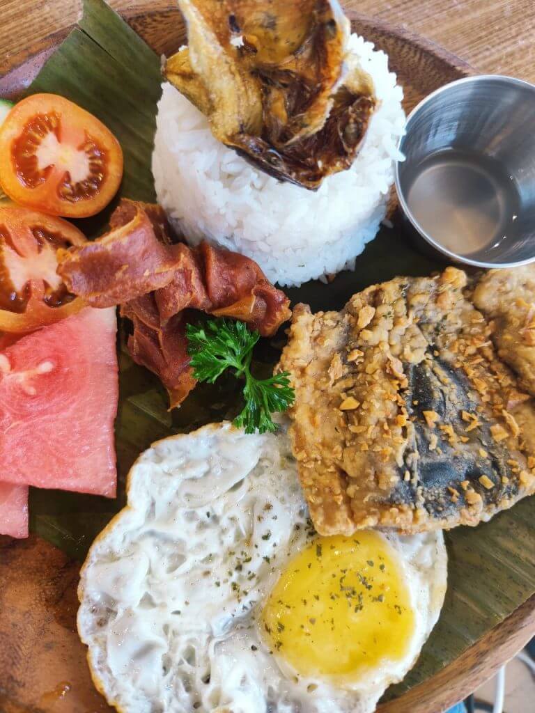 a plate with rice, fried egg, fish, bacon, tomato &amp; watermelon at urban coffee studio&rsquo;s coffee shop.