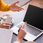 two people discuss freelancing opportunities at a table with a laptop, coffee, and phone in a cozy setting.