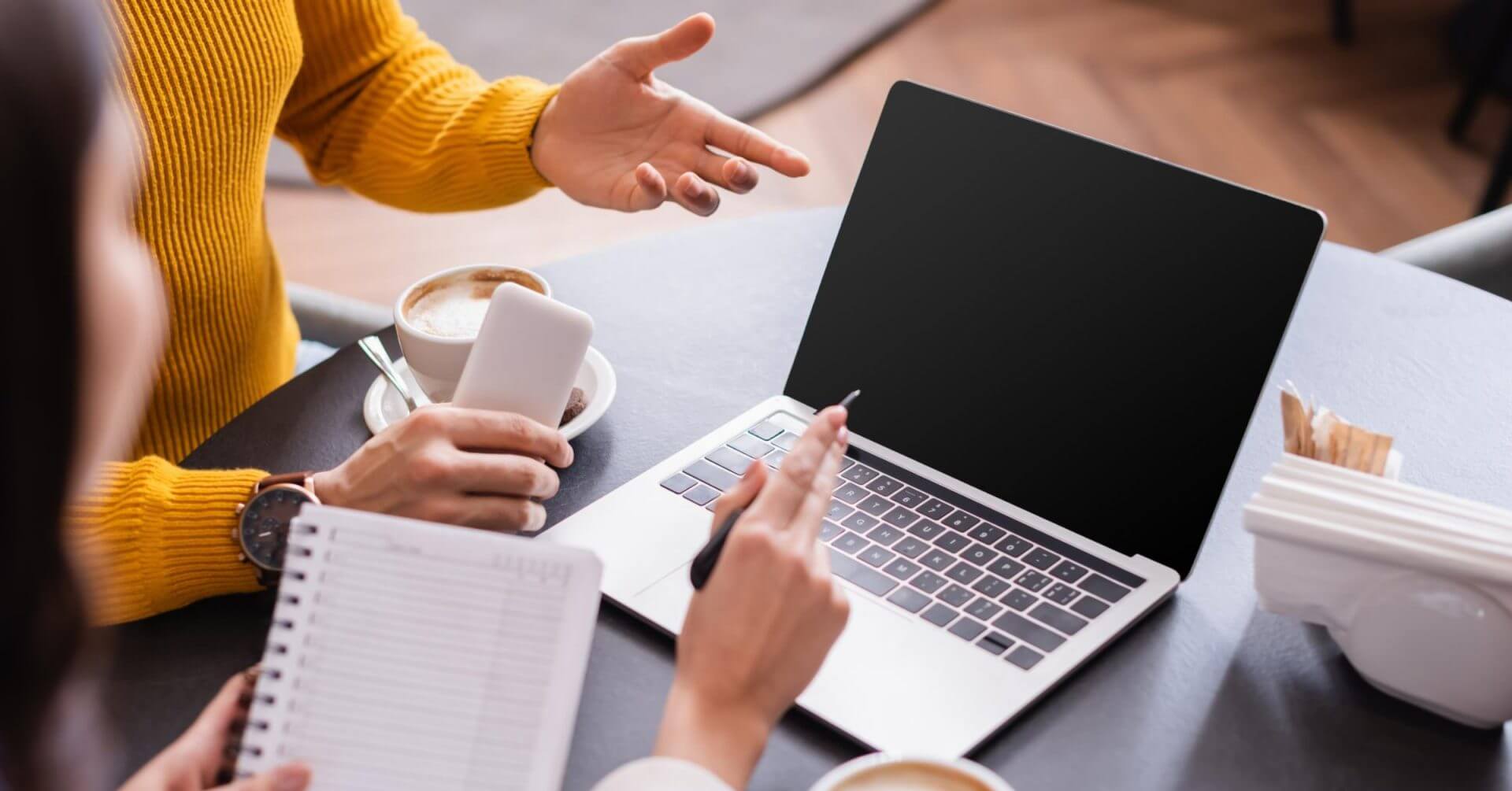 two people discuss freelancing opportunities at a table with a laptop, coffee, and phone in a cozy setting.
