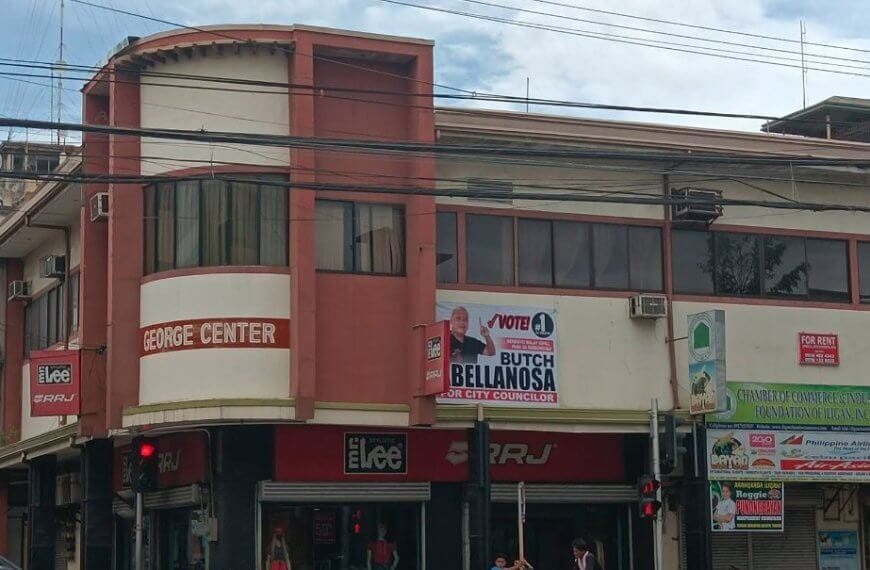 a two story commercial real estate building with shops, signs, and a political campaign banner on the corner.