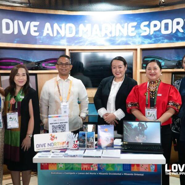 six people stand smiling at a dive and marine sports booth with brochures, laptop, and colorful display.