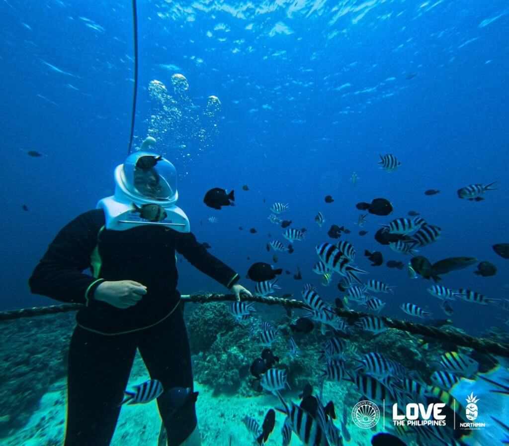 a person in a sea helmet dives among camiguin&rsquo;s fish; sunken cemetery and love the philippines logos visible.