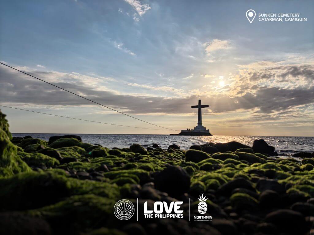 a large cross rises from the sea at sunset; moss covered rocks line the sunken cemetery shore in camiguin.